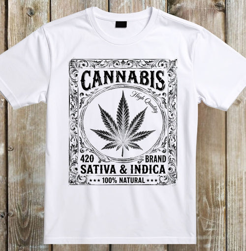 A white short-sleeve crewneck t-shirt with a black graphic print on the front, featuring a cannabis leaf and decorative text, displayed flat against a rustic wooden plank background.