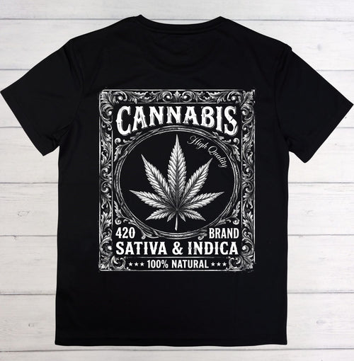 A black short-sleeve crewneck t-shirt laid flat on a white wood plank background. The front features a large white graphic print styled like a vintage ornate label, containing a cannabis leaf, the word 'CANNABIS', 'High Quality', '420 BRAND', 'SATIVA & INDICA', and '100% NATURAL'.