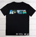 A black short-sleeve t-shirt laid flat on a white wood plank background. The front features a graphic spelling 'NOLA' in stylized letters filled with scenic images of New Orleans, and the text 'HEY YOU, SEE ME,' is printed in white near the bottom hem.