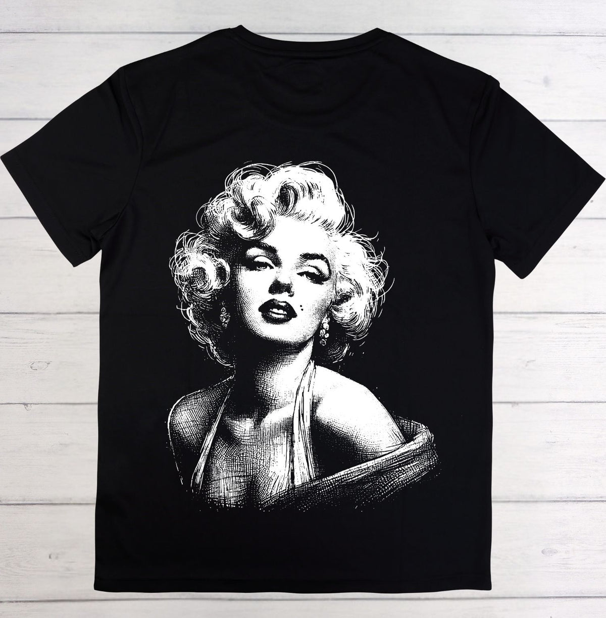 A black short-sleeve t-shirt featuring a large white graphic print of Marilyn Monroe on the front, laid flat on a light-colored wooden plank background.