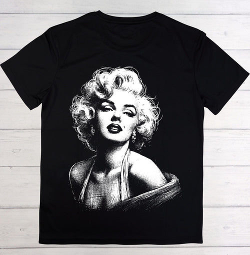 A black short-sleeve t-shirt featuring a large white graphic print of Marilyn Monroe on the front, laid flat on a light-colored wooden plank background.