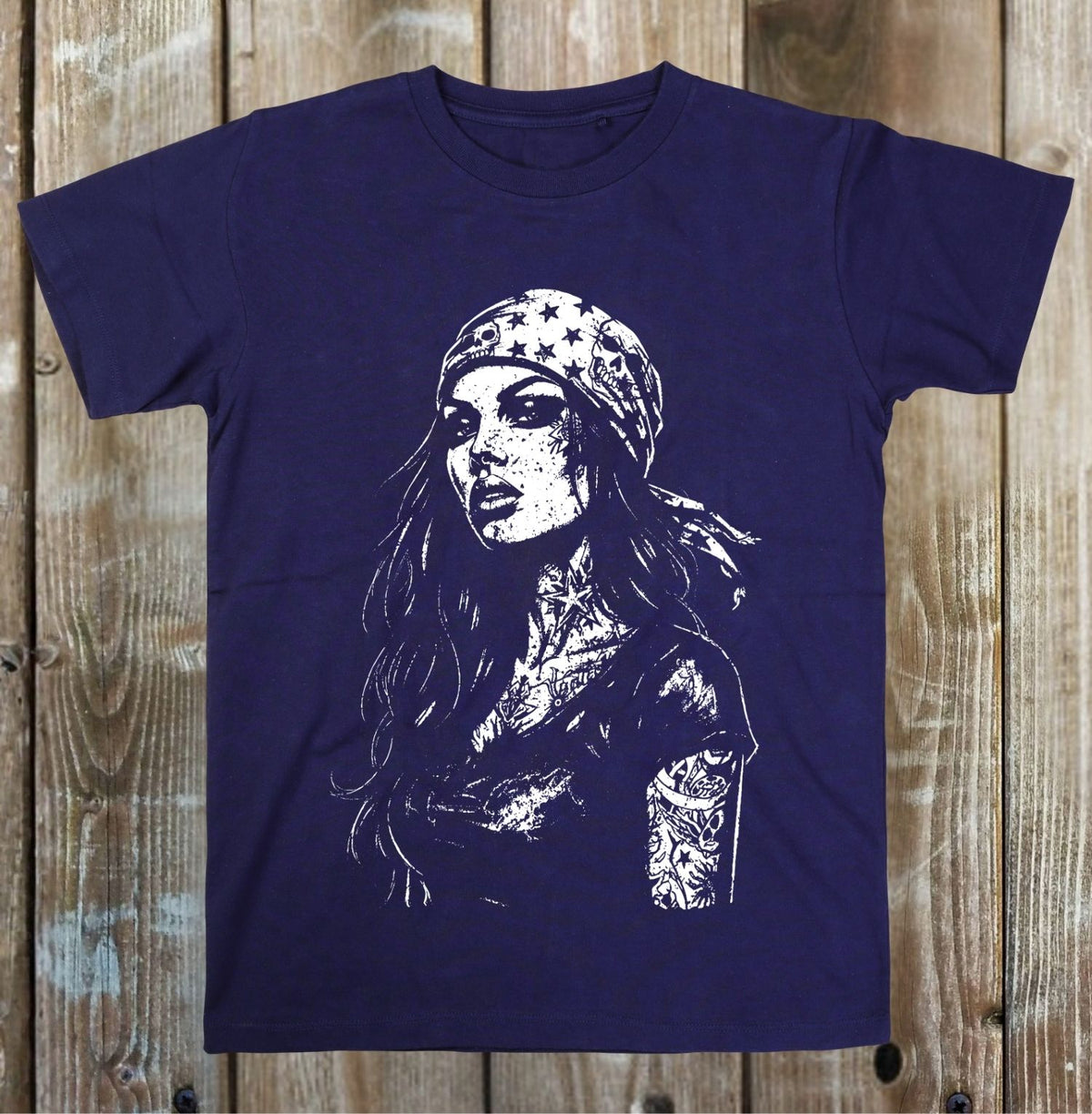 A navy blue short-sleeve t-shirt with a white graphic print of a tattooed woman wearing a bandana, displayed flat against a rustic wooden plank background.