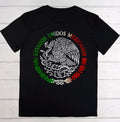 A black short-sleeve t-shirt laid flat on a white wood plank background, featuring a large circular Mexican emblem graphic on the chest with the words 'ESTADOS UNIDOS MEXICANOS' in green, white, and red.
