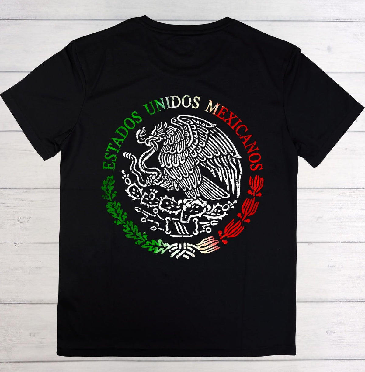 A black short-sleeve t-shirt laid flat on a white wood plank background, featuring a large circular Mexican emblem graphic on the chest with the words 'ESTADOS UNIDOS MEXICANOS' in green, white, and red.