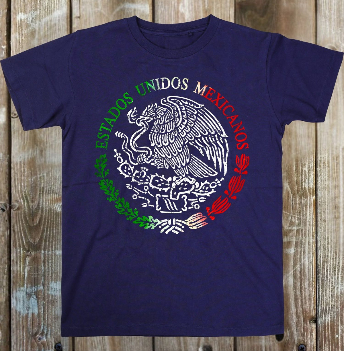 A navy blue short-sleeve t-shirt with a circular graphic of the Mexican coat of arms in the center, surrounded by the text 'ESTADOS UNIDOS MEXICANOS' in green, white, and red, laid flat on a rustic wooden plank background.