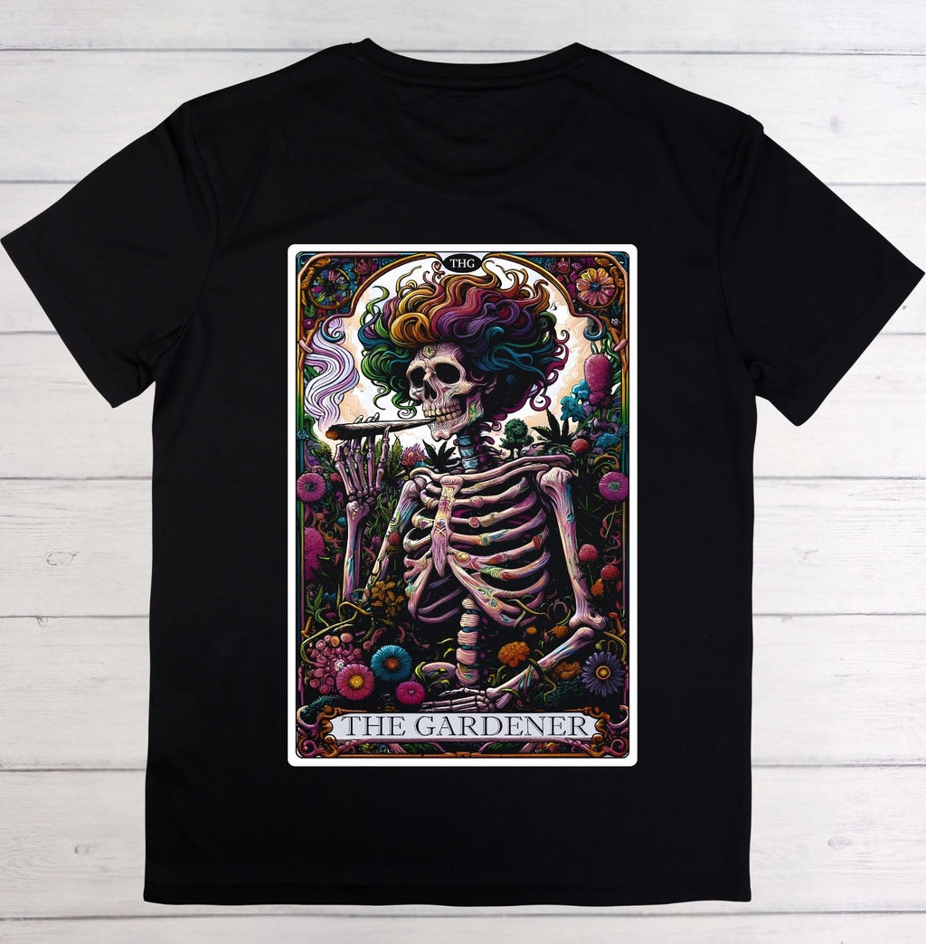 A black short-sleeve crewneck t-shirt featuring a large graphic print of 'The Gardener' tarot card on the front, displayed flat against a light gray wood plank background.