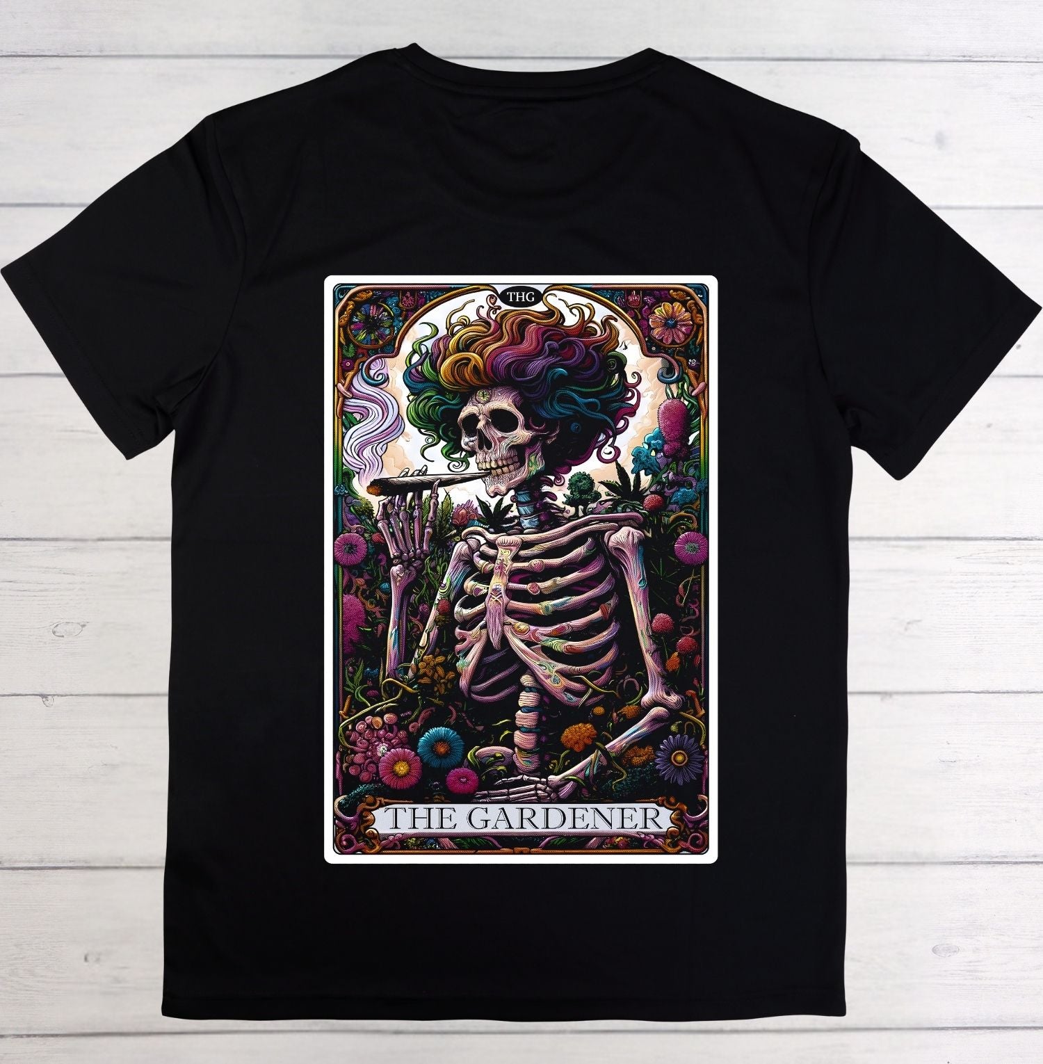 A black short-sleeve crewneck t-shirt featuring a large graphic print of 'The Gardener' tarot card on the front, displayed flat against a light gray wood plank background.