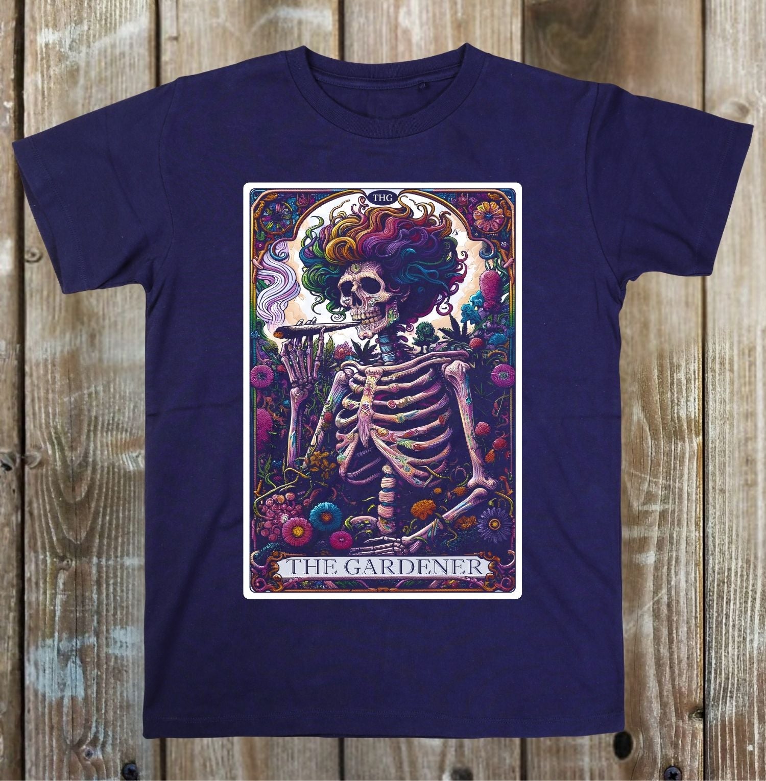 A navy blue short-sleeve t-shirt with a colorful graphic print of a skeleton holding a cigar, styled like 'The Gardener' tarot card, displayed flat against a rustic wooden plank background.