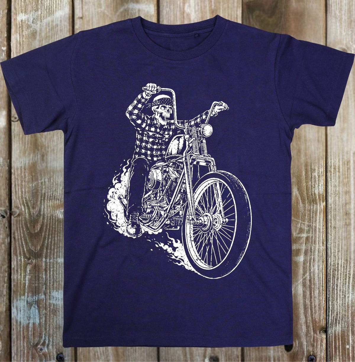 A navy blue short-sleeve t-shirt with a white graphic print of a skeleton riding a chopper motorcycle, displayed flat against a rustic wooden plank background.