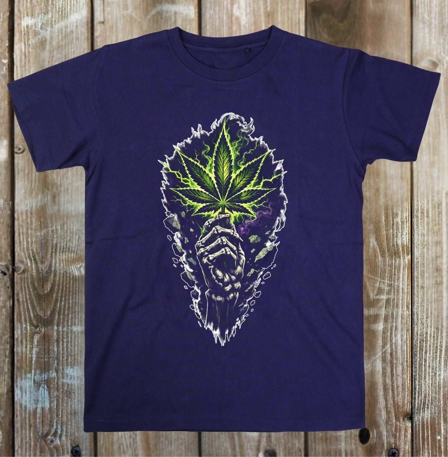 A navy blue short-sleeve t-shirt with a graphic print of a green cannabis leaf on a white skull, laid flat on a rustic wooden plank background.