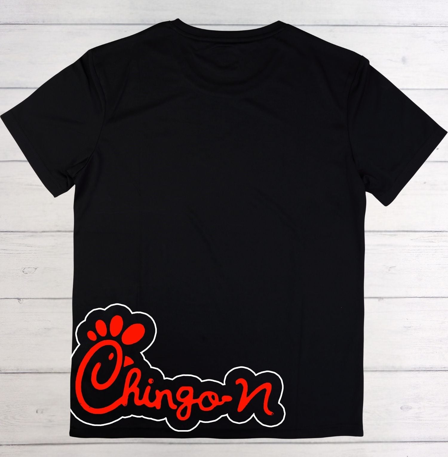 A black short-sleeve t-shirt laid flat on a white wood plank background, featuring a red and white 'Chingo-N' graphic with a paw print outline near the lower left front.