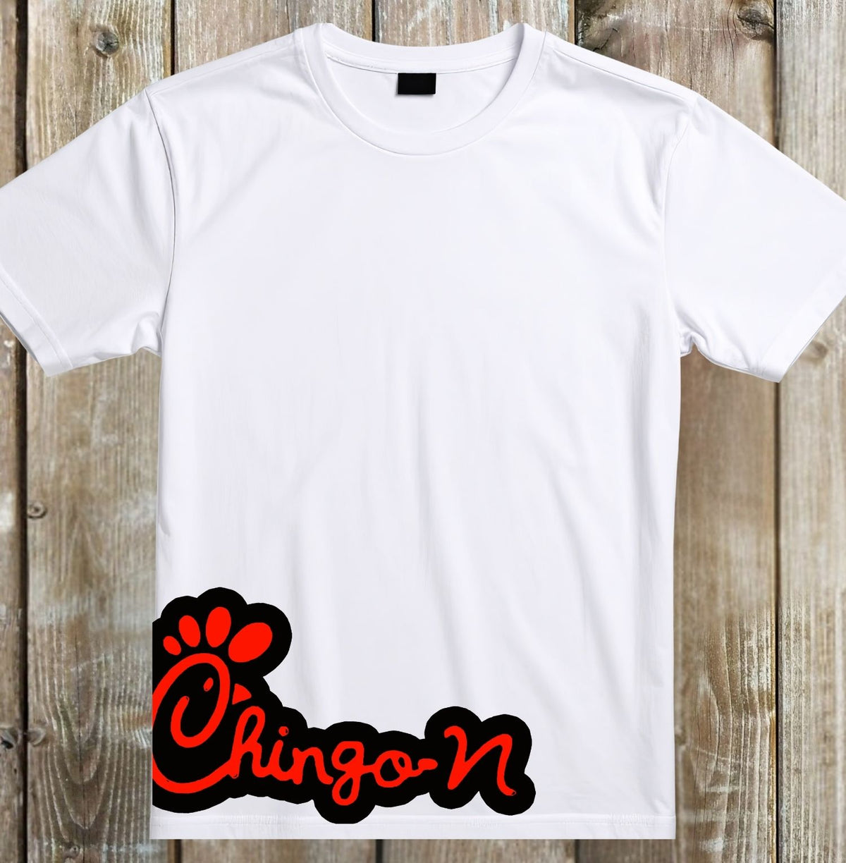 A white short-sleeve crewneck t-shirt laid flat on a rustic wooden plank background, featuring a red and black graphic on the lower left that reads 'Chingo' with a paw print.