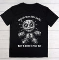 A black short-sleeve t-shirt with a white graphic of a stitched voodoo doll and the text 'CROSS MY HEART, HOPE YOU DIE' and 'Stick A Needle in Your Eye', displayed flat against a rustic wooden plank background.