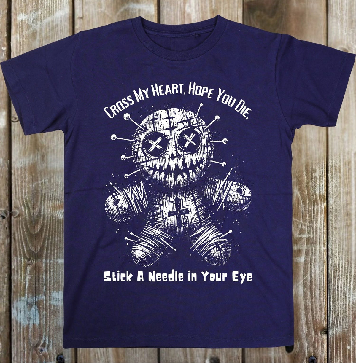 A navy blue short-sleeve t-shirt with a white graphic of a stitched voodoo doll and the text 'CROSS MY HEART, HOPE YOU DIE' and 'Stick A Needle in Your Eye', displayed flat against a rustic wooden plank background.