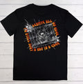 A black short-sleeve graphic t-shirt laid flat on a white wood plank background. The front of the shirt features a large graphic print with a white and gray bear inside a cage, surrounded by orange and yellow flame-style text that reads 'DEBUTTE ALL MY DAYS A MAT TO A CAGE'.