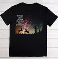 A black short-sleeve t-shirt with a graphic print on the front, laid flat on a white wood plank background. The graphic features silhouettes of two jumping deer, a tree, and a galaxy sky with the text 'PUSHING LITTLE CHILDREN WITH THEIR FULLY AUTOMATIC'.