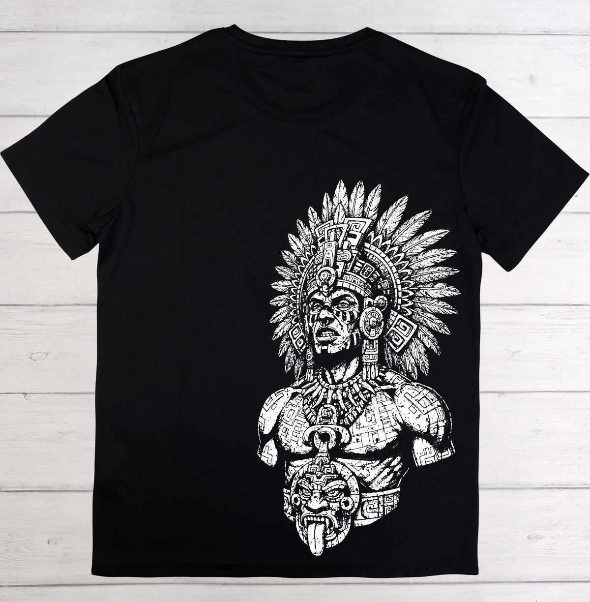 A black short-sleeve t-shirt with a large white graphic print of a detailed Aztec-style warrior wearing a feathered headdress, laid flat on a white wood plank background.