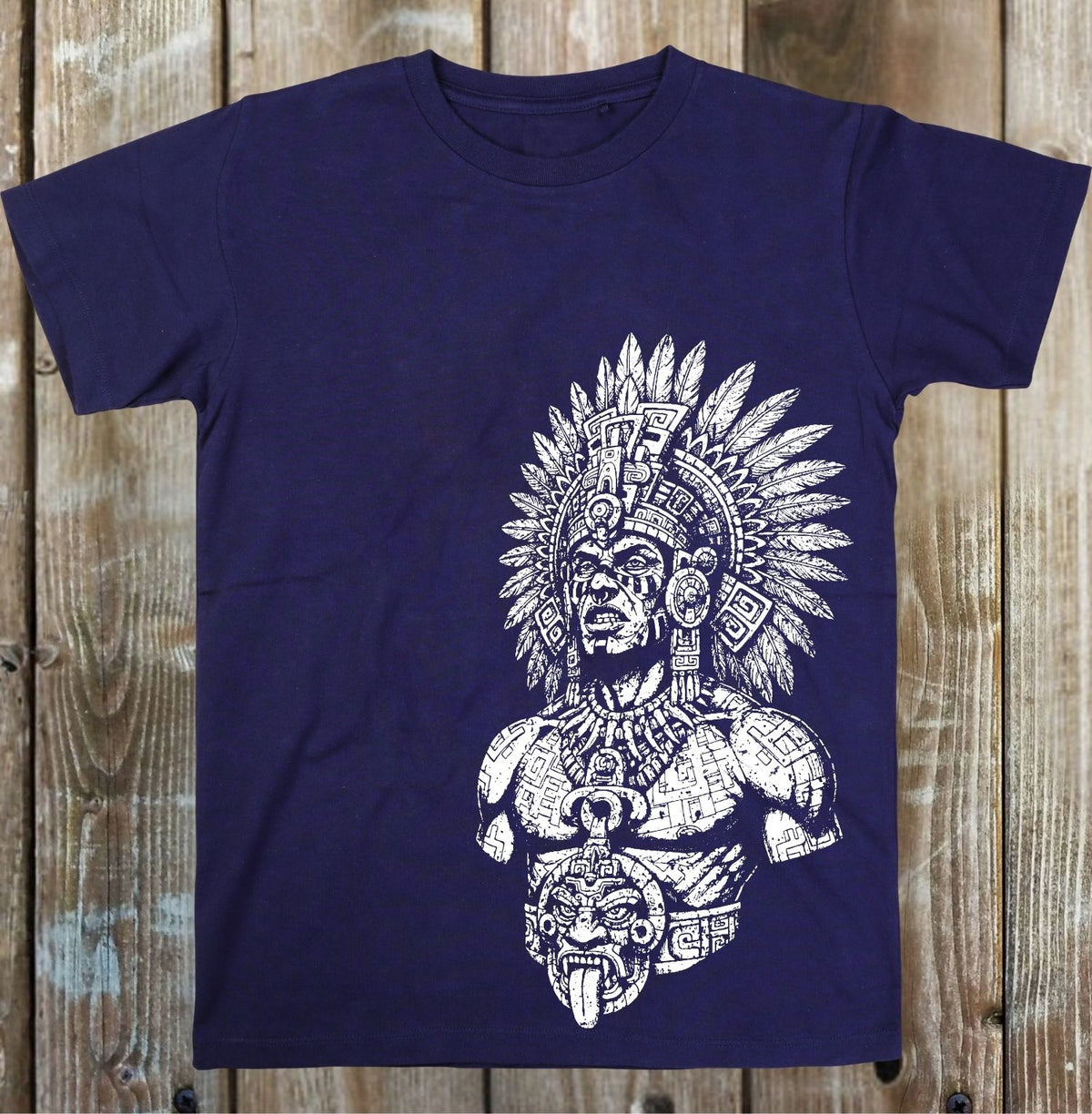 A navy blue short-sleeve t-shirt with a large white graphic print of an Aztec-style warrior wearing a detailed feathered headdress, laid flat on a rustic wooden plank background.