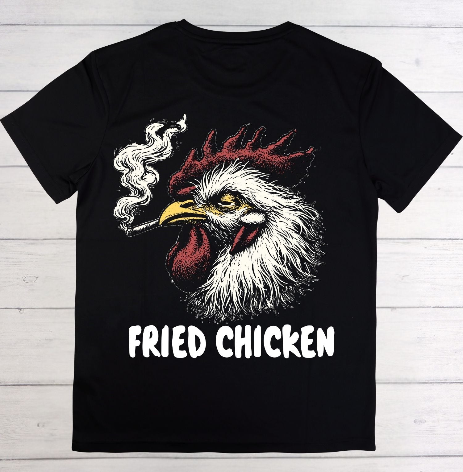 A black short-sleeve t-shirt laid flat on a white wood plank background, featuring a graphic of a smoking rooster with the words 'FRIED CHICKEN' printed below in white.