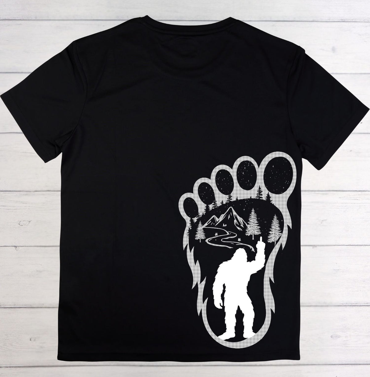 A black short-sleeve t-shirt laid flat on a white wood plank background, featuring a large white graphic on the front with a bear paw print enclosing a mountain landscape and a silhouetted figure.