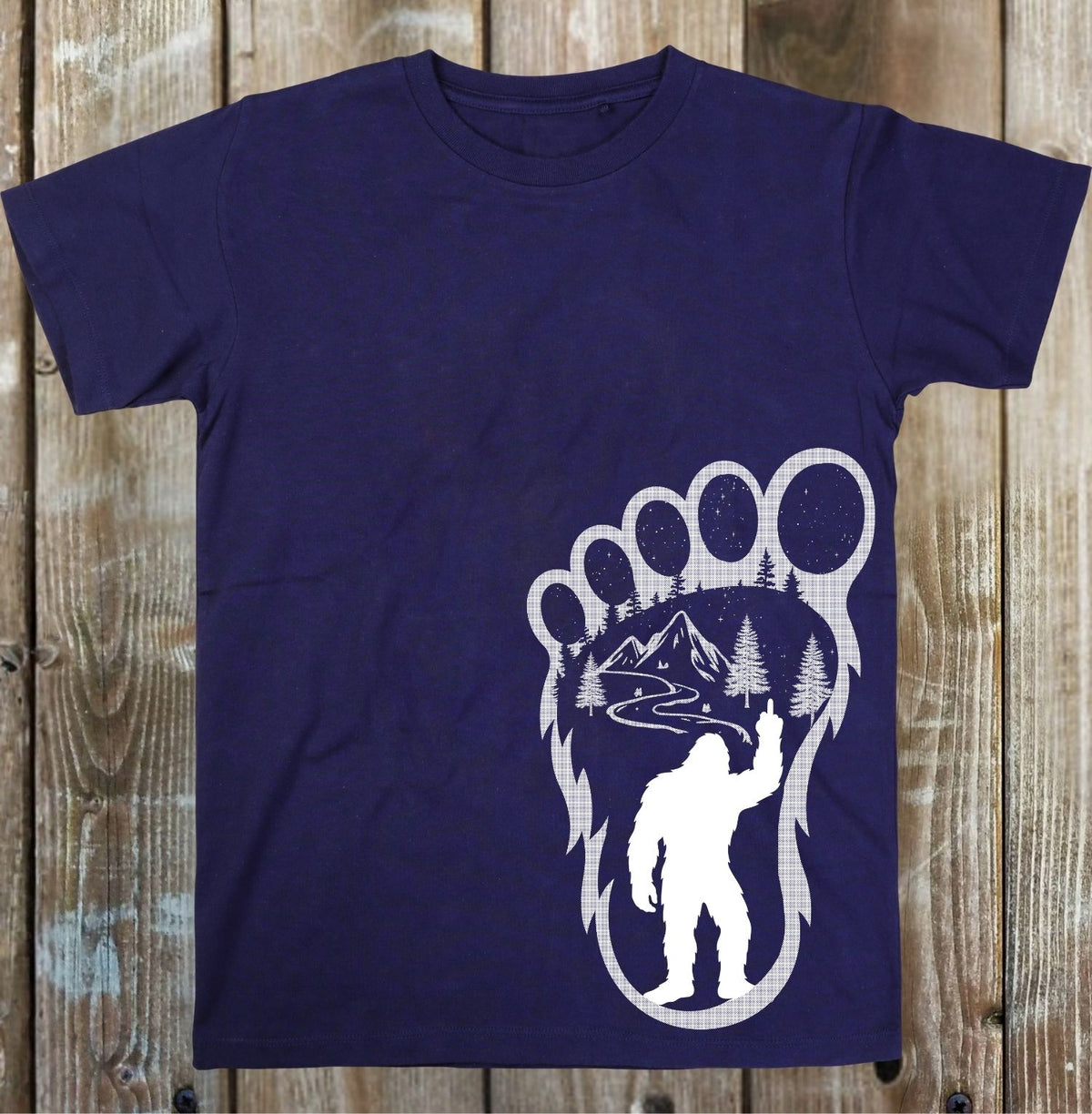A navy blue short-sleeve t-shirt with a white graphic print of a Bigfoot silhouette inside a giant bear paw footprint containing a mountain and river scene, displayed flat against a rustic wooden plank background.