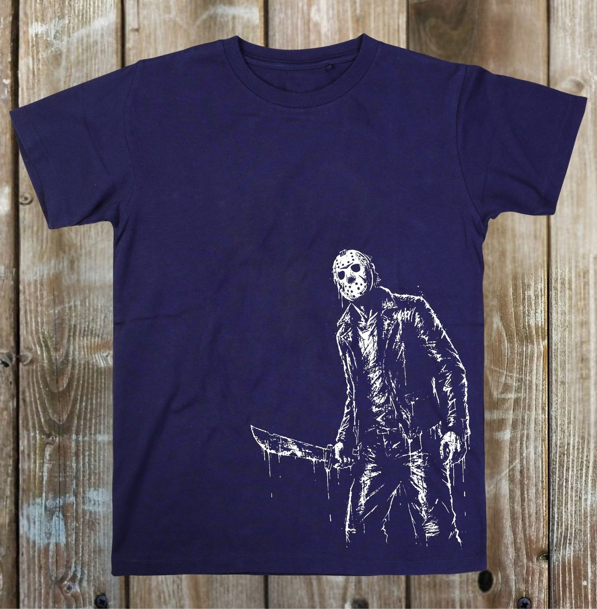 A dark navy blue short-sleeve t-shirt with a white distressed graphic of Jason Voorhees holding a knife, displayed flat against a rustic wooden plank background.