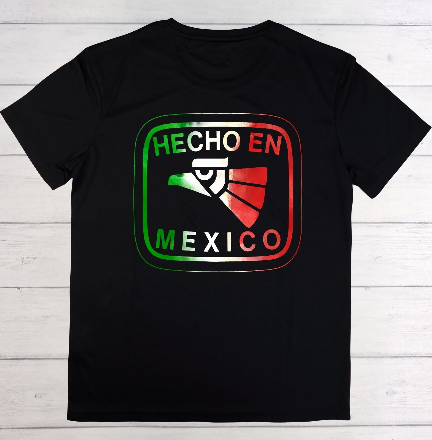 A black short-sleeve t-shirt laid flat on a white wood plank background, featuring a large graphic on the chest with the text 'HECHO EN MEXICO' and a stylized eagle in green, white, and red.