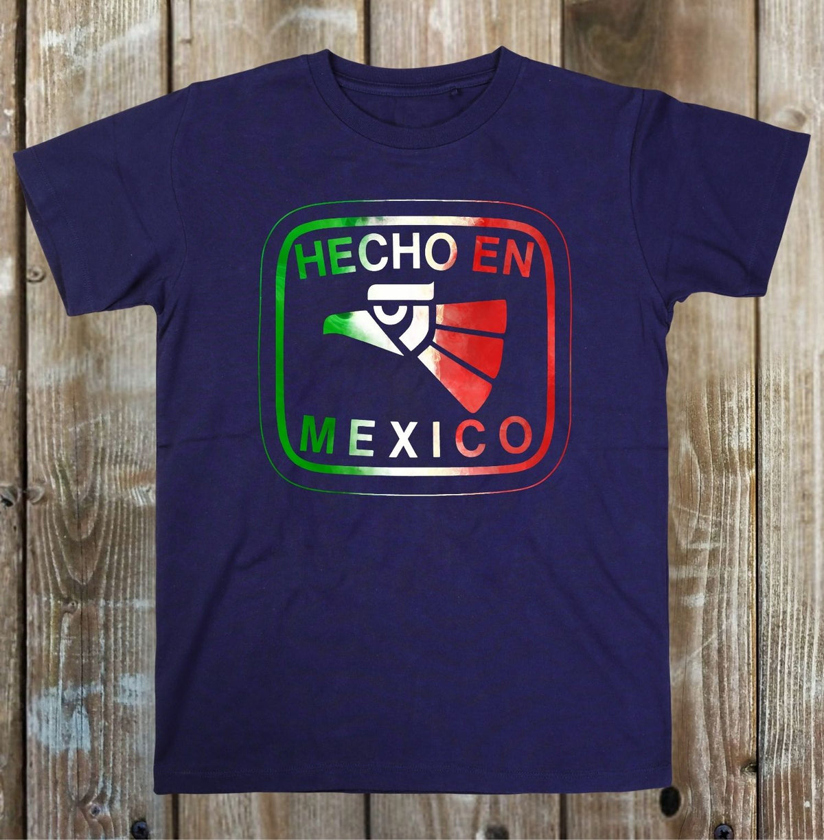 A navy blue short-sleeve t-shirt with a graphic on the chest that reads 'HECHO EN MEXICO' in white, outlined in green and red, with a Mexican flag motif. The t-shirt is laid flat on a rustic wooden plank background.