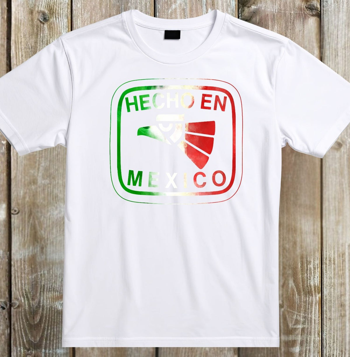 A white short-sleeve crewneck t-shirt laid flat on a rustic wooden plank background. The front of the shirt features a graphic print with the text 'HECHO EN MEXICO' in white, layered over a green-to-white-to-red gradient badge containing a green and white eagle emblem.