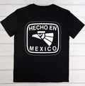A black short-sleeve t-shirt with a white graphic print on the front that reads 'HECHO EN MEXICO' around an eagle head illustration, laid flat on a light wood plank background.