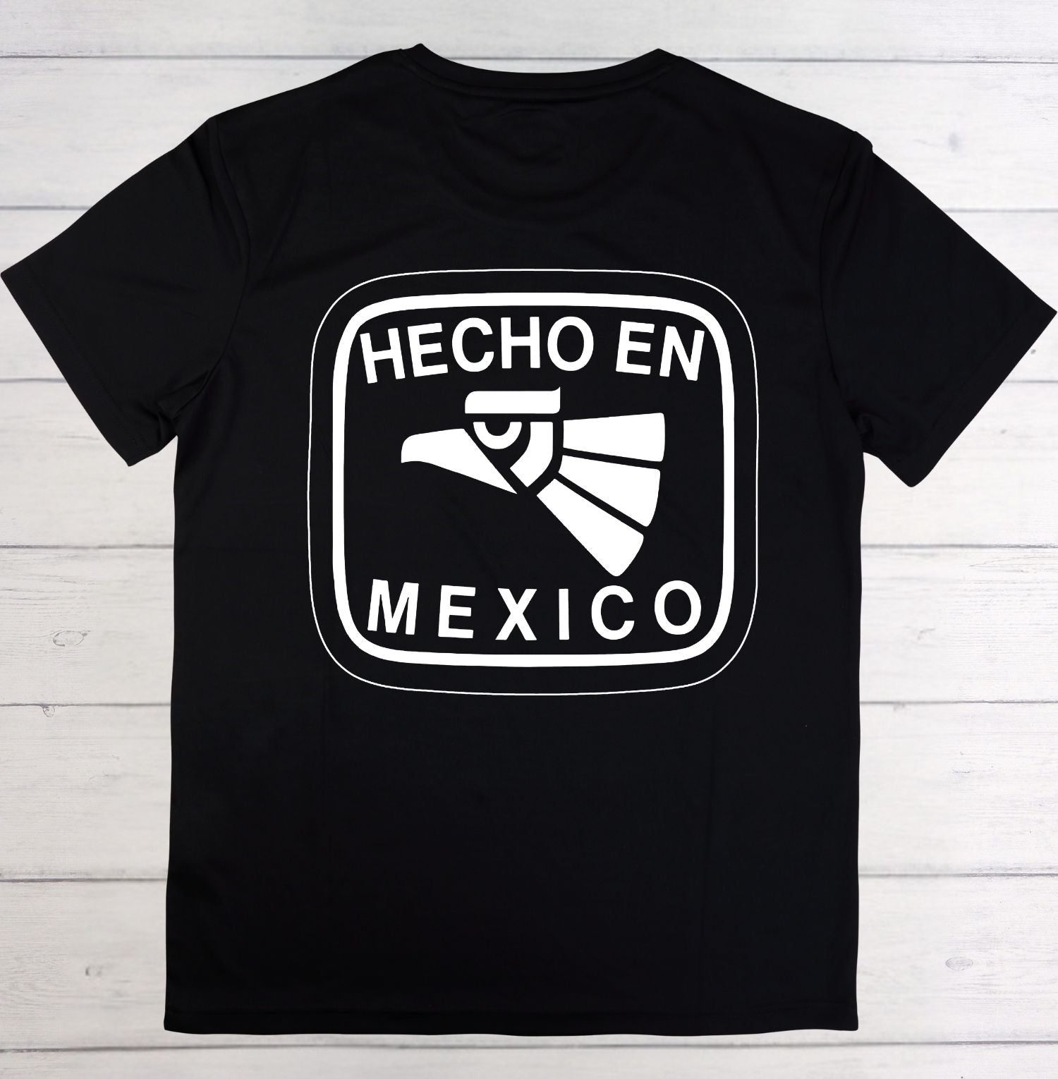 A black short-sleeve t-shirt with a white graphic print on the front that reads 'HECHO EN MEXICO' around an eagle head illustration, laid flat on a light wood plank background.