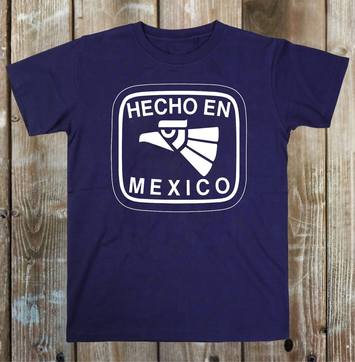 A navy blue short-sleeve t-shirt with a white graphic print on the front that reads 'HECHO EN MEXICO' around an eagle head illustration, displayed flat against a rustic wooden plank background.