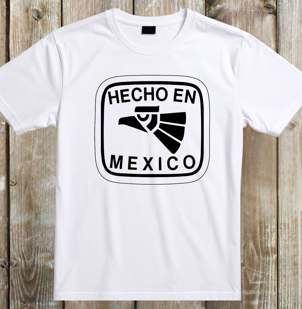 A white short-sleeve crewneck t-shirt with a black graphic print on the chest reading 'HECHO EN MEXICO' around a stylized eagle head, laid flat on a rustic wooden plank background.