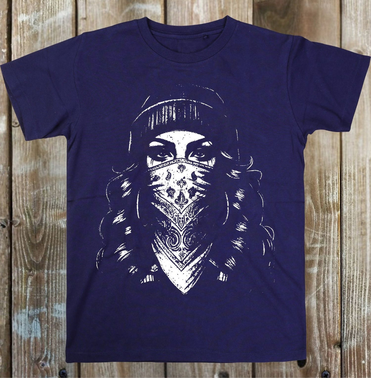 A navy blue short-sleeve t-shirt with a white graphic print of a woman wearing a patterned bandana and beanie, displayed flat against a rustic wooden plank background.