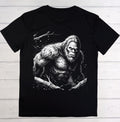 A black short-sleeve crewneck t-shirt featuring a detailed black and white graphic print of a Bigfoot figure on the front, laid flat on a light-colored wooden plank background.