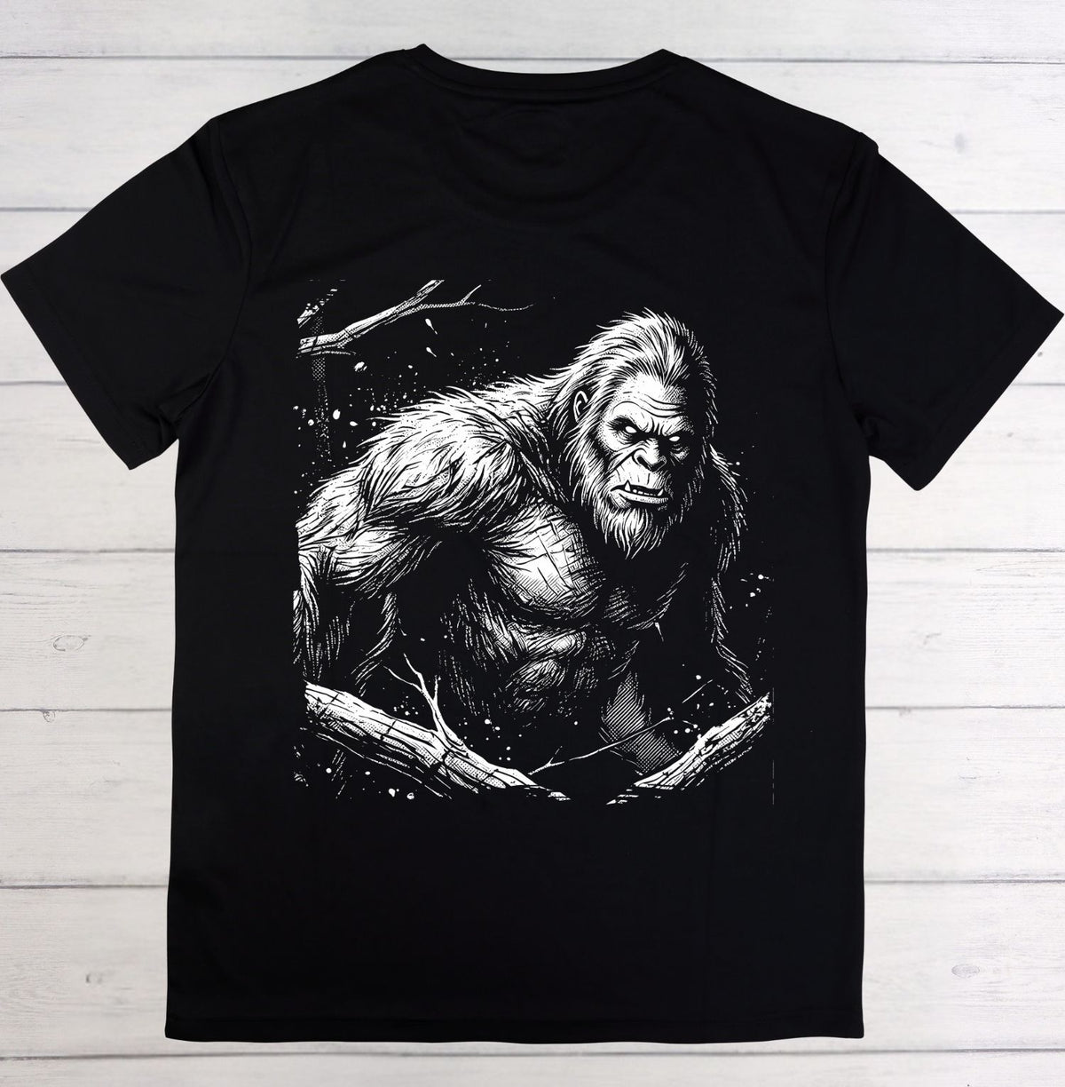 A black short-sleeve crewneck t-shirt featuring a detailed black and white graphic print of a Bigfoot figure on the front, laid flat on a light-colored wooden plank background.