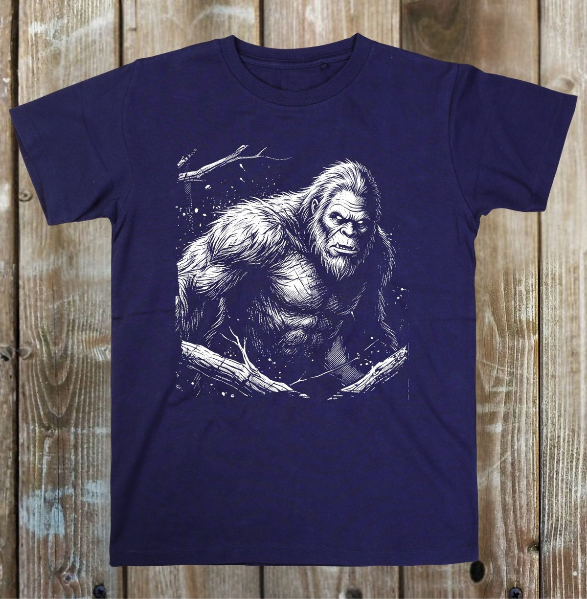 A navy blue short-sleeve t-shirt with a detailed white graphic of Bigfoot, displayed flat against a rustic wooden plank background.