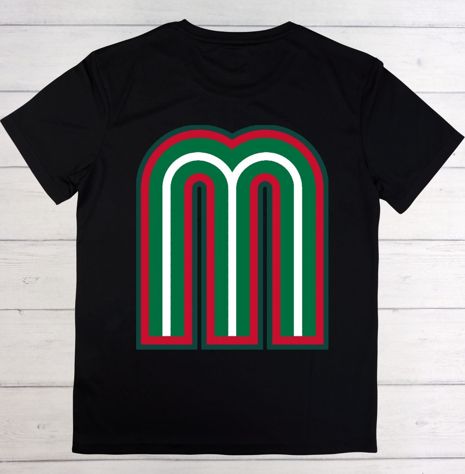A black short-sleeve t-shirt laid flat on a white wood plank background, featuring a large stylized letter 'M' graphic on the chest with concentric arches in green, red, and white.