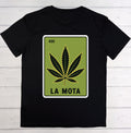 A black short-sleeve t-shirt laid flat on a white wood plank background, featuring a graphic print of a green card with a black cannabis leaf and the text 'LA MOTA' and '420'.