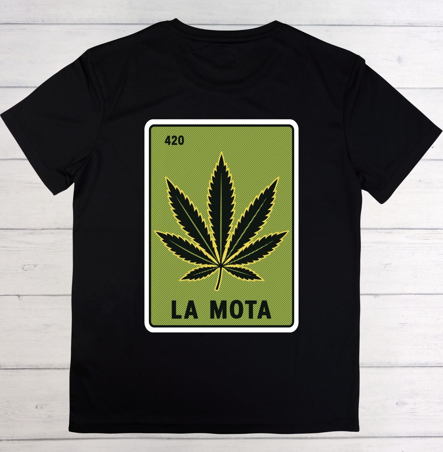 A black short-sleeve t-shirt laid flat on a white wood plank background, featuring a graphic print of a green card with a black cannabis leaf and the text 'LA MOTA' and '420'.