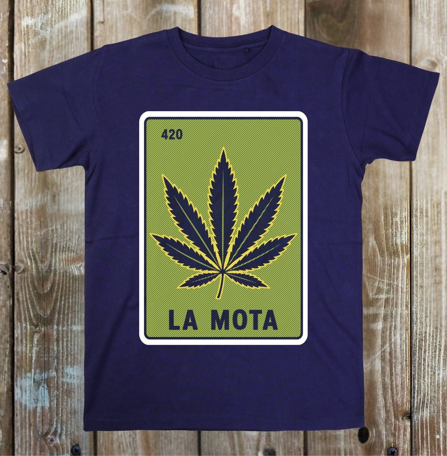 A navy blue short-sleeve t-shirt with a graphic print of a cannabis leaf styled like a green highway sign reading 'LA MOTA' and '420', displayed flat against a rustic wooden plank background.