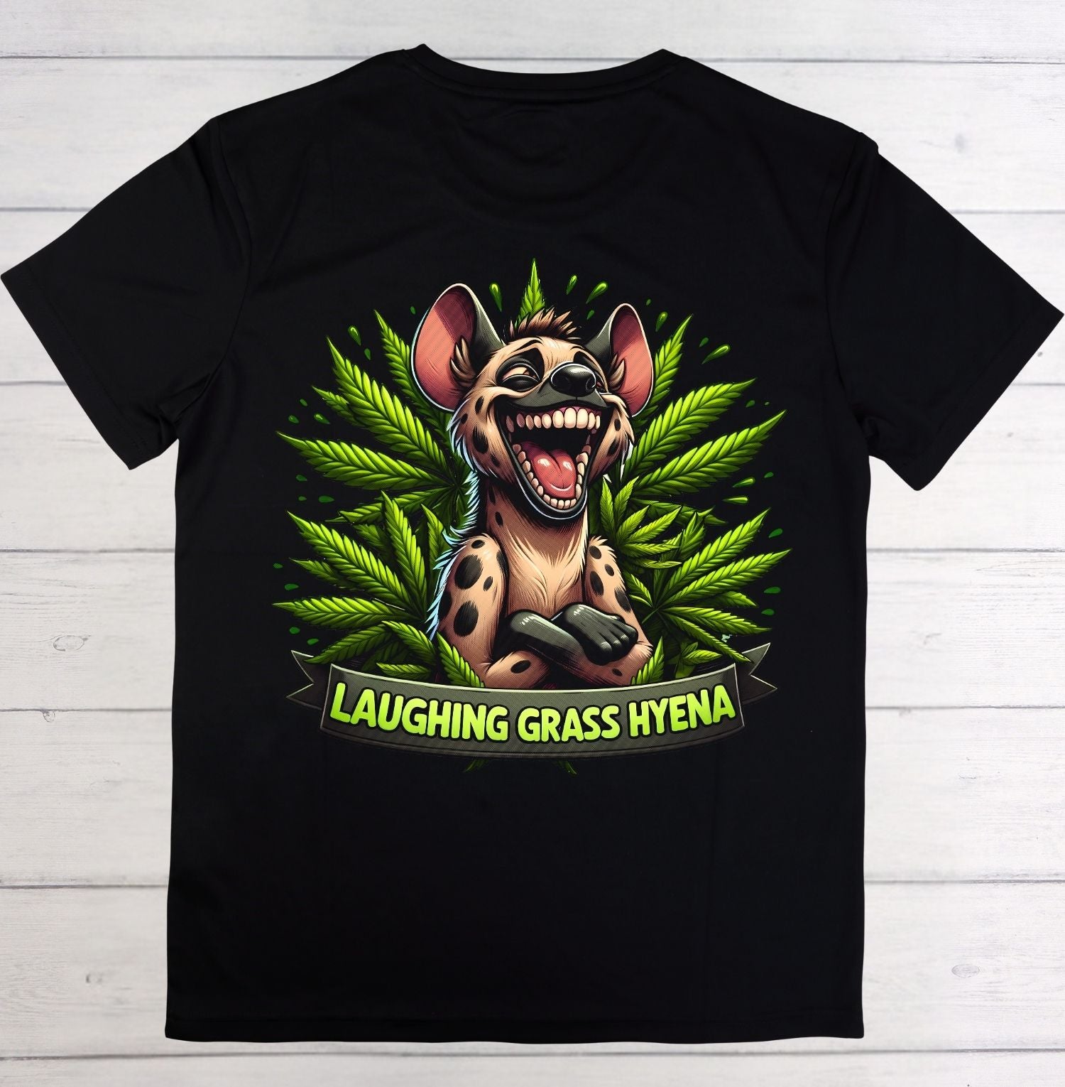 A black short-sleeve t-shirt laid flat on a white wood background, featuring a colorful graphic of a laughing grass hyena surrounded by green leaves, with a banner reading 'LAUGHING GRASS HYENA'.