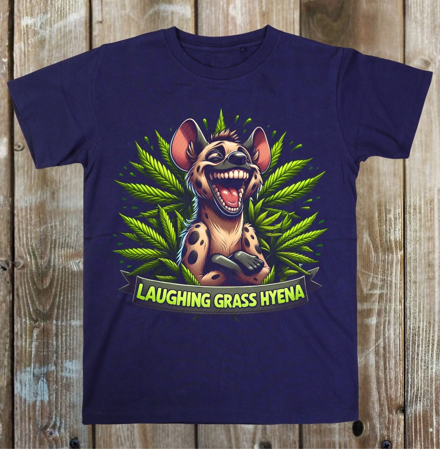 A navy blue short-sleeve t-shirt with a colorful graphic of a laughing grass hyena surrounded by green cannabis leaves, displayed flat against a rustic wooden plank background.