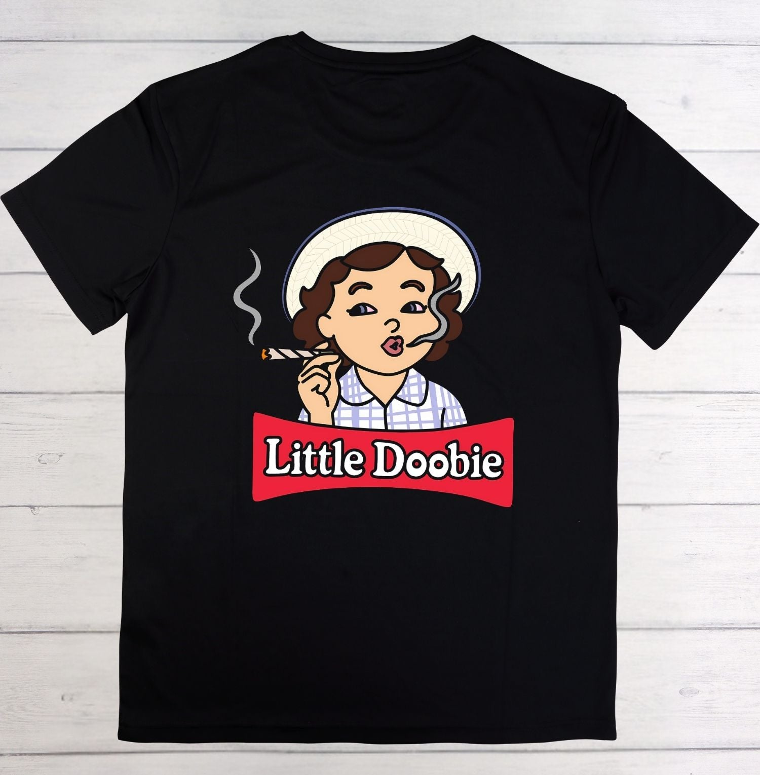 A black short-sleeve t-shirt laid flat on a white wood plank background, featuring a cartoon graphic of a woman with brown hair, a white hat, and a red banner that reads 'Little Doobie'.