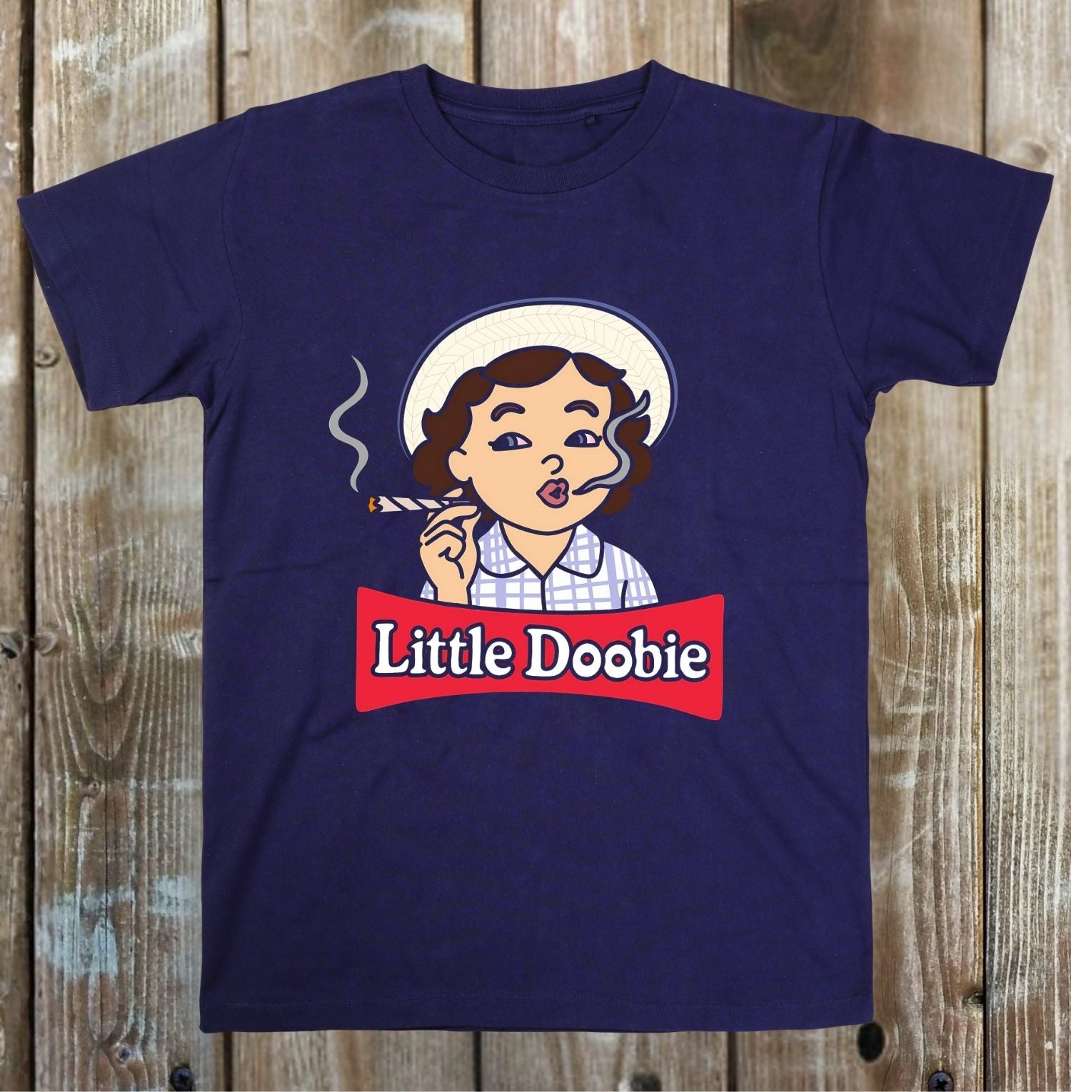 A navy blue short-sleeve t-shirt with a cartoon graphic of a woman smoking a cigarette, with a red banner reading 'Little Doobie' underneath, displayed flat against a rustic wooden plank background.