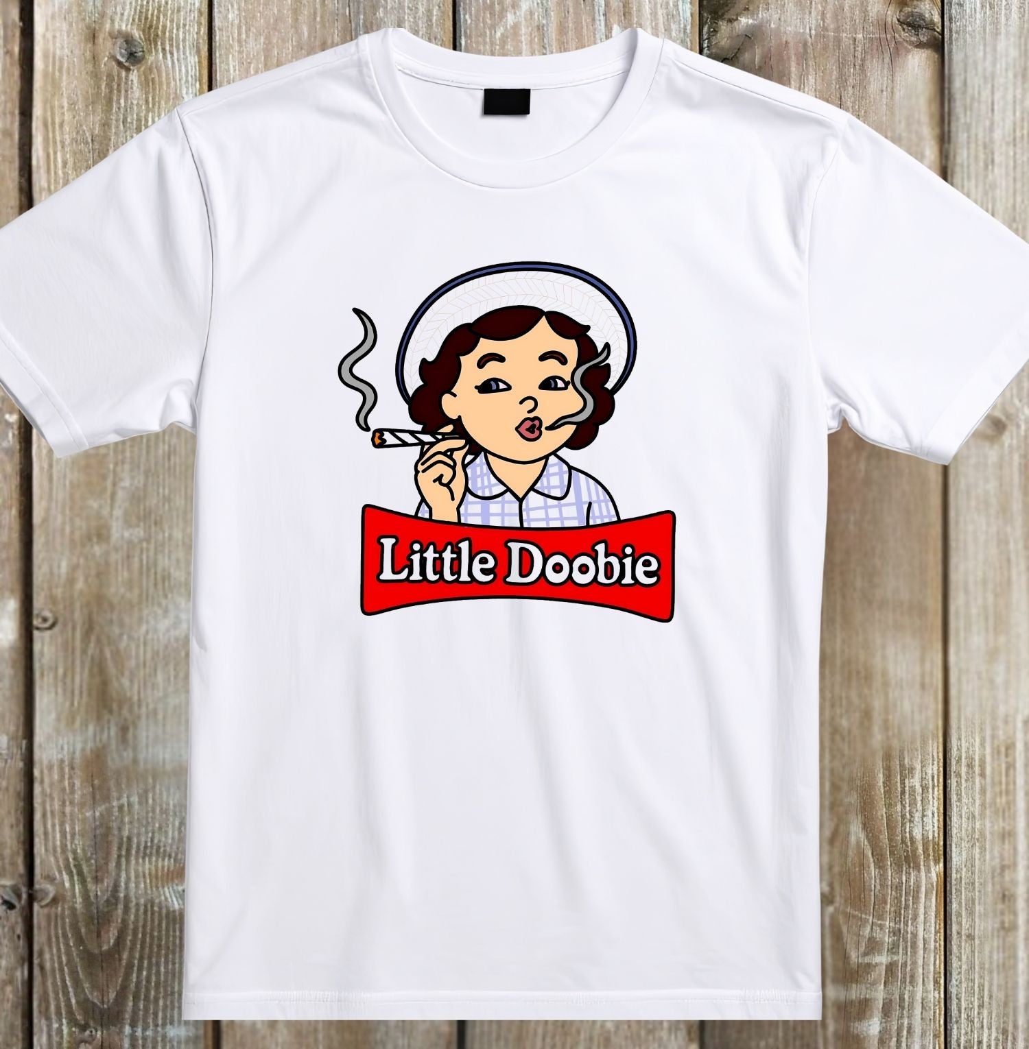 A white short-sleeve crewneck t-shirt laid flat on a rustic wooden background, featuring a cartoon graphic of a woman smoking with a red banner that reads 'Little Doobie'.