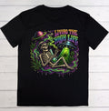 A black short-sleeve t-shirt laid flat on a white wood background, featuring a colorful graphic of a skeleton holding a green bong with a green liquid, a UFO, a planet, and the text 'LIVING THE HIGH LIFE' in neon green, purple, and pink.