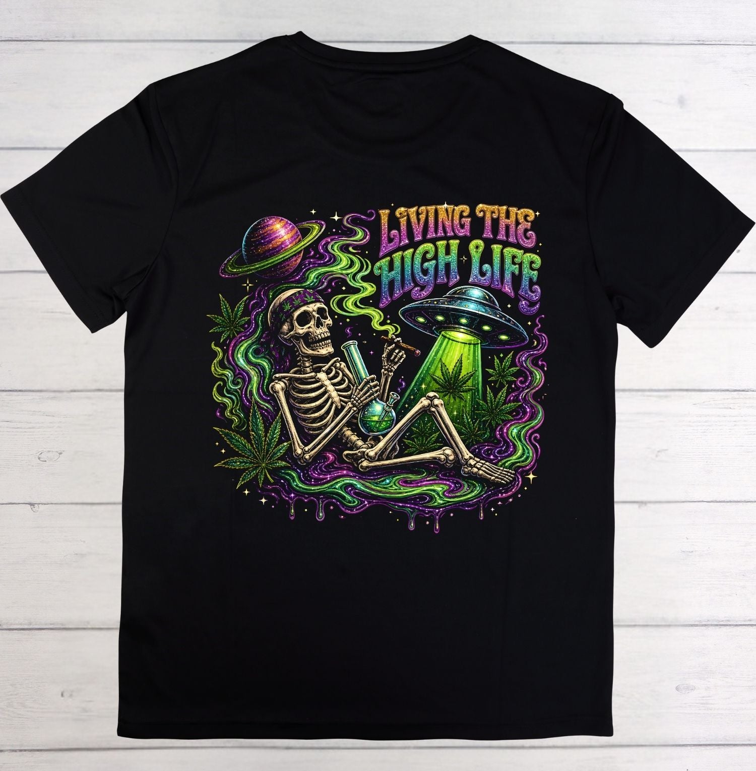 A black short-sleeve t-shirt laid flat on a white wood background, featuring a colorful graphic of a skeleton holding a green bong with a green liquid, a UFO, a planet, and the text 'LIVING THE HIGH LIFE' in neon green, purple, and pink.