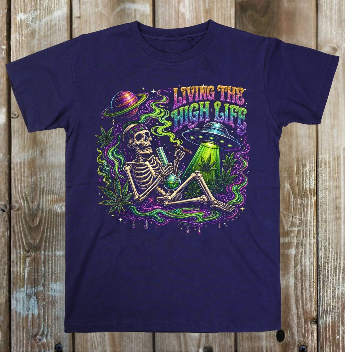 A navy blue short-sleeve t-shirt with a colorful retro graphic of a skeleton sitting next to a green bong and a UFO, with the text 'LIVING THE HIGH LIFE' in a rainbow gradient. The t-shirt is displayed flat against a rustic wooden plank background.