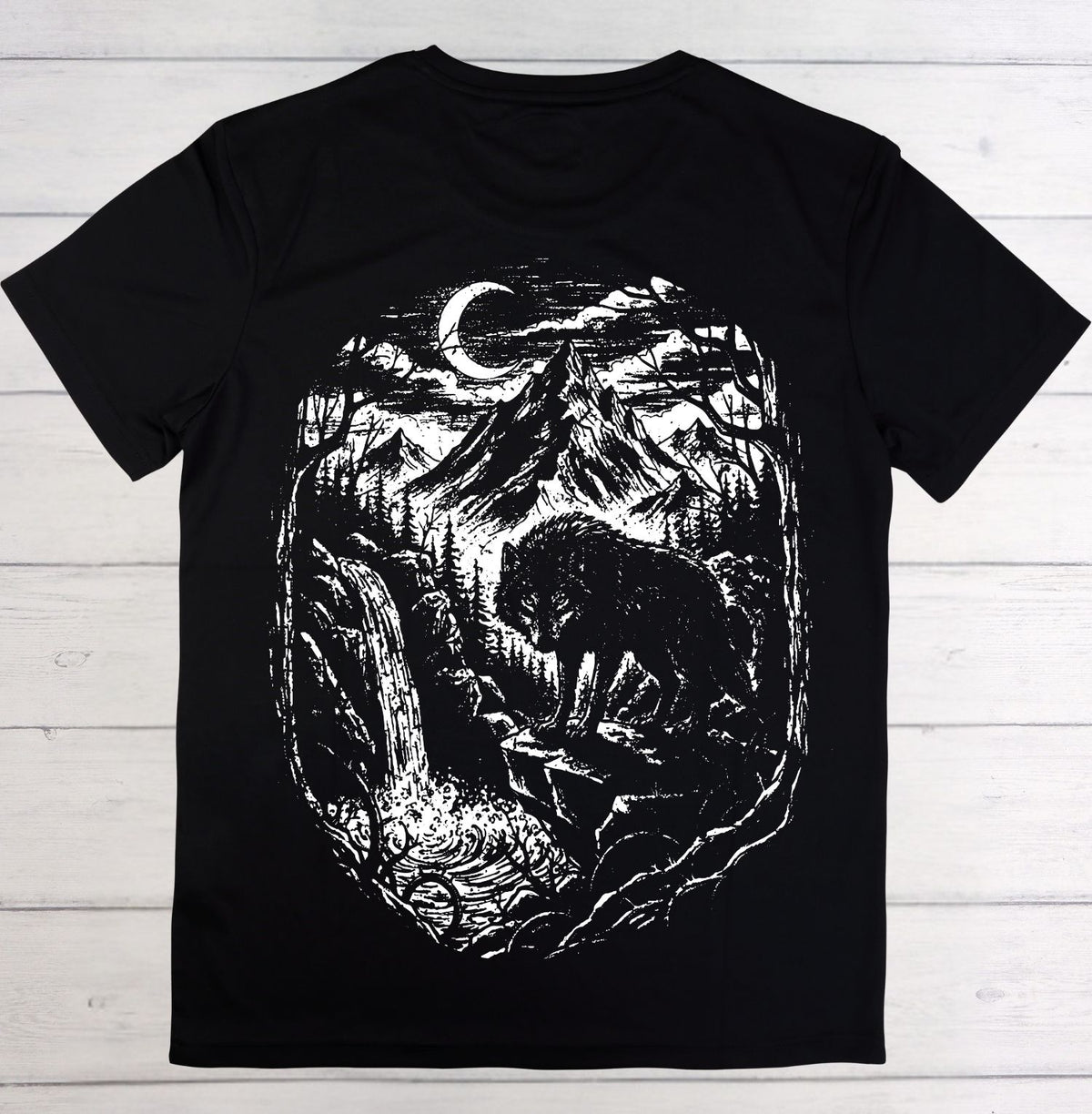 A black short-sleeve t-shirt laid flat on a white wood plank background, featuring a large white graphic print of a wolf in a forest under a crescent moon.